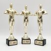 Trys aukso spalvos žmogaus figūros statulėlės su nominacijų lentelėmis ant juodų pagrindų.