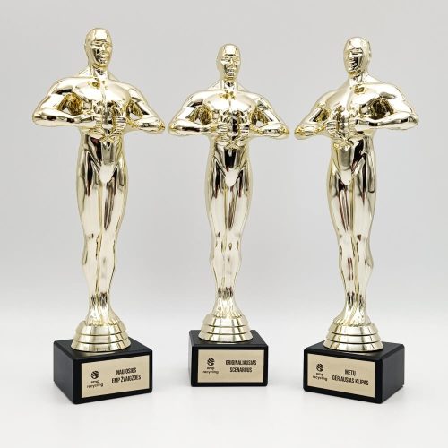 Trys aukso spalvos žmogaus figūros statulėlės su nominacijų lentelėmis ant juodų pagrindų.