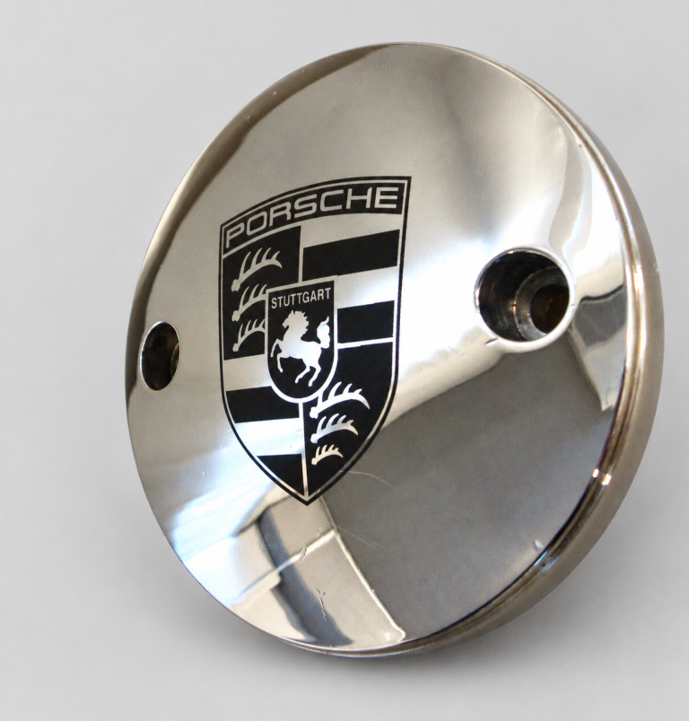 Blizgi metalinė detalė su juodu „Porsche“ herbu šviesiai pilkame fone